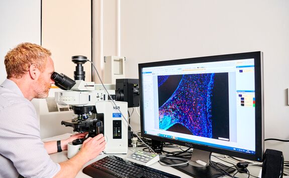 Person using a microscope while viewing a colourful microscopic image of cellular structures on a computer screen. The desk holds various lab tools and equipment.
